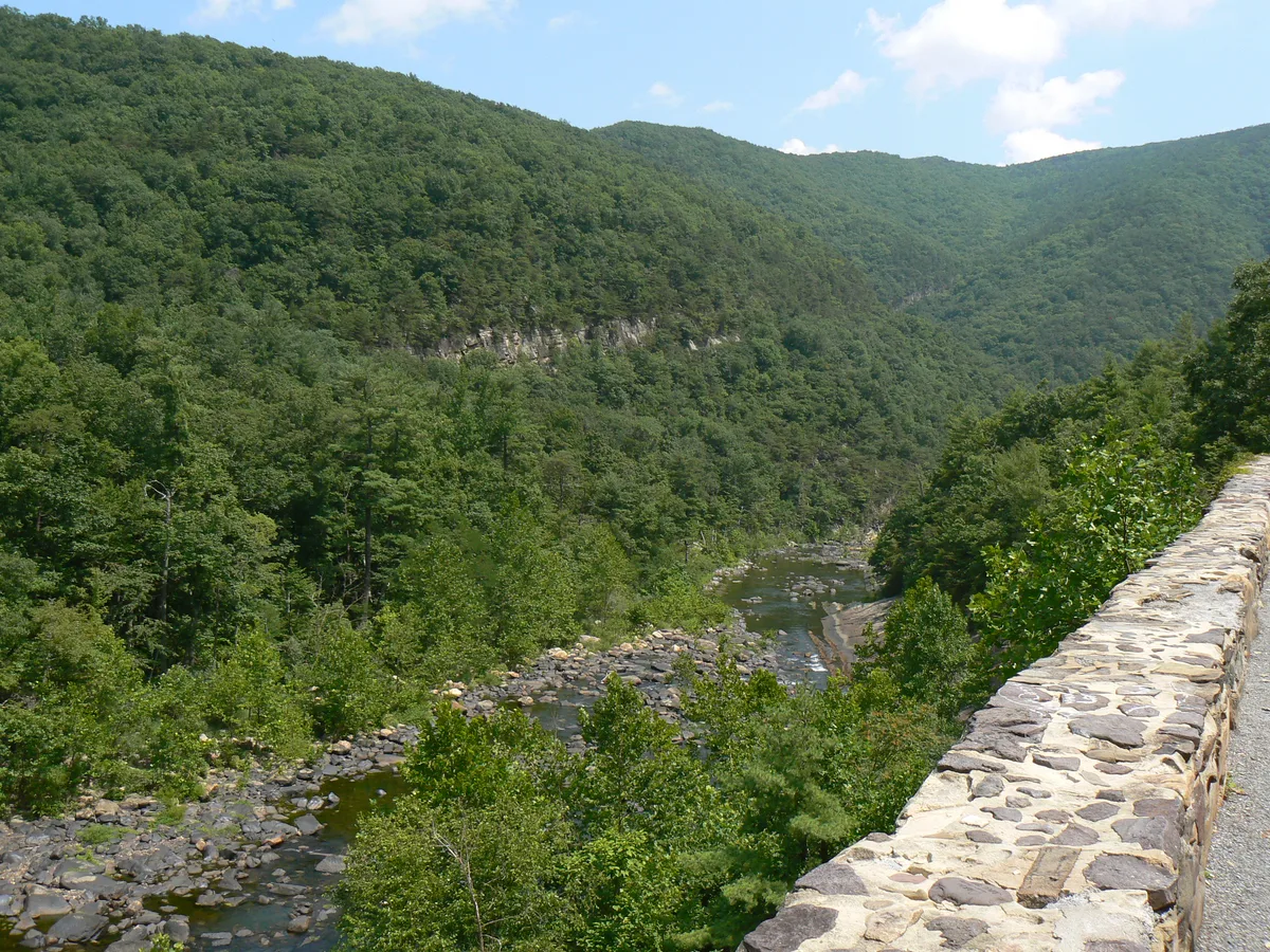 The Maury River winds through a deep, forested gorge in Goshen Pass, with rocky shoals and boulders below steep green mountains. A stone guardwall lines the overlook at right beneath a bright blue morning sky with scattered clouds.