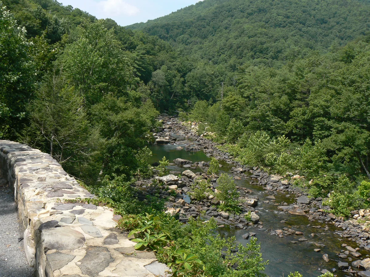 A rocky river winds through a steep, forested gorge, seen from a stone guardwall along the overlook. Dense green mountains rise on both sides, with the Maury River broken by boulders and small riffles below.