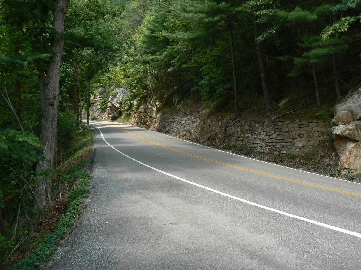 A winding two-lane road curves through a narrow, wooded mountain pass, with a stone retaining wall and exposed rock outcrops lining the right side. Tall evergreens and dense summer foliage frame the scene in soft morning light.