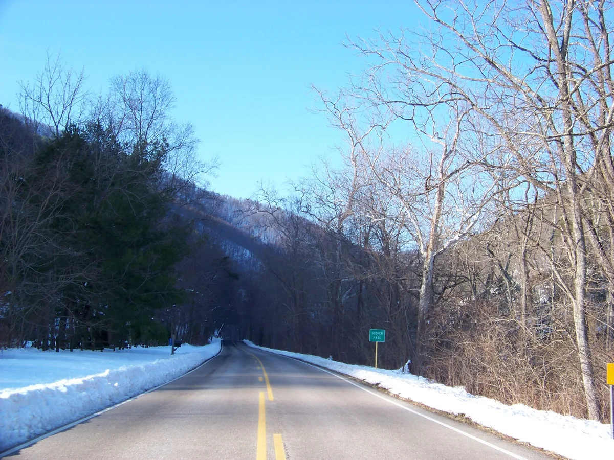 A two-lane road curves into a narrow, tree-lined mountain pass under a clear blue sky, with snow piled along both shoulders and bare winter branches arching over the pavement. A small green “Goshen Pass” sign marks the roadside near the wooded gorge.