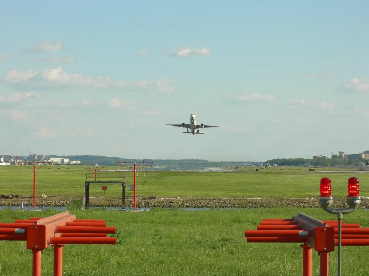 A jet approaches low over the Potomac under a pale blue sky, framed by Gravelly Point’s orange benches and runway lights in the foreground. The wide grassy field and distant airport buildings emphasize the plane’s near-overhead landing path.