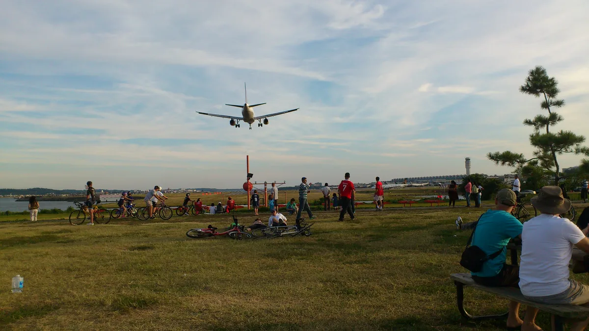 A commercial jet descends low over Gravelly Point Park, centered against a wide pastel sky as spectators watch from the grassy riverfront. Bicycles, picnic tables, and the Reagan National control tower frame the scene in warm late-day light.