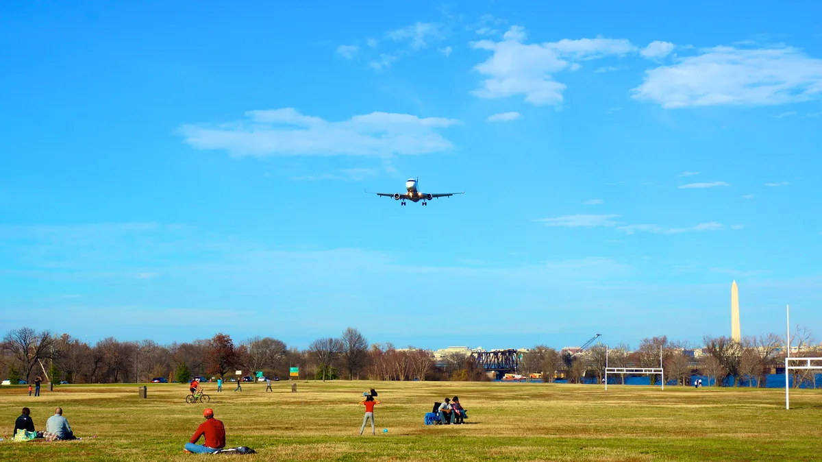 A jet on final approach flies low over Gravelly Point’s open grassy field, with people picnicking and watching beneath a bright blue sky. The Washington Monument rises in the distance beyond the Potomac, adding a clean skyline backdrop to the wide scene.