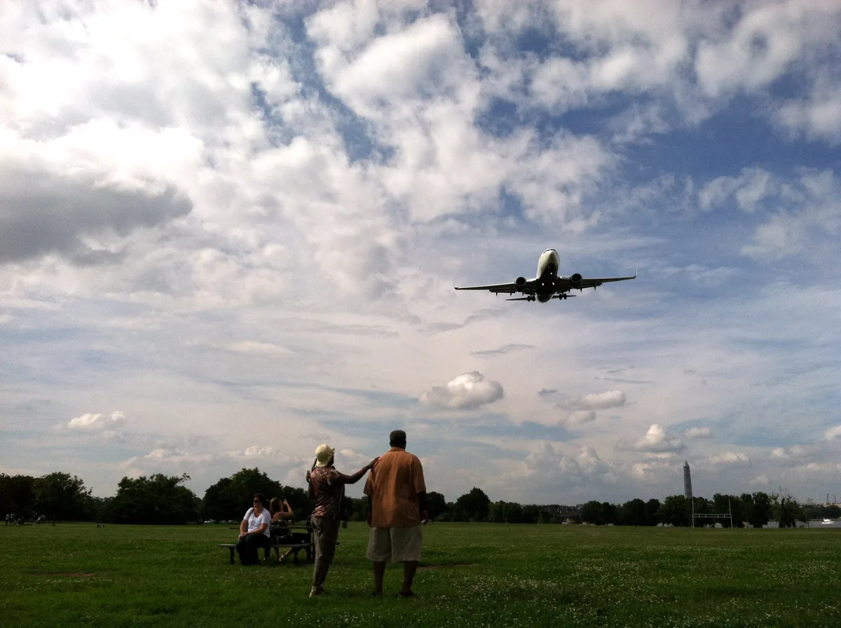 A low-flying passenger jet approaches overhead against a dramatic, cloud-filled sky above Gravelly Point’s wide green field. In the foreground, a couple stands watching near a picnic table as the Potomac-side park stretches toward a distant Washington Monument.