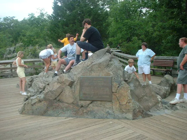Visitors gather around a large natural boulder with a bronze plaque mounted on its face, set in the middle of a wooden boardwalk overlook. Trees and railings frame the scene, with people sitting, standing, and climbing on the rock in a casual summer setting.