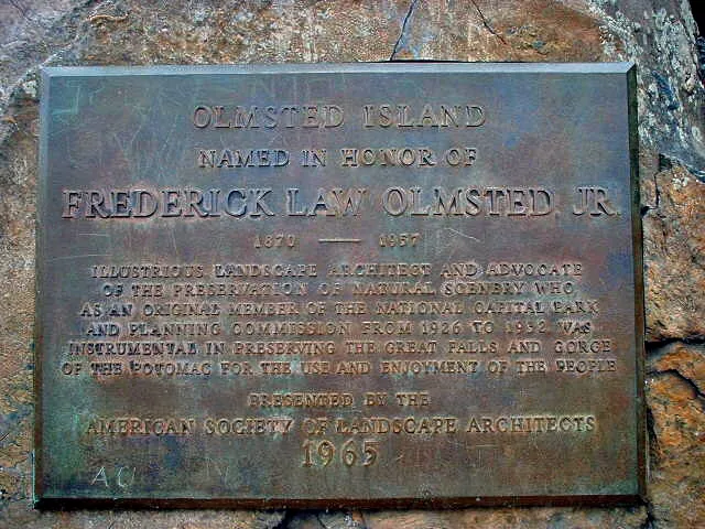 A weathered bronze plaque mounted on rough stone reads “Olmsted Island” and honors Frederick Law Olmsted Jr., with engraved text centered in raised lettering. The plaque’s dark patina and rock backdrop give the scene a historic, understated feel.