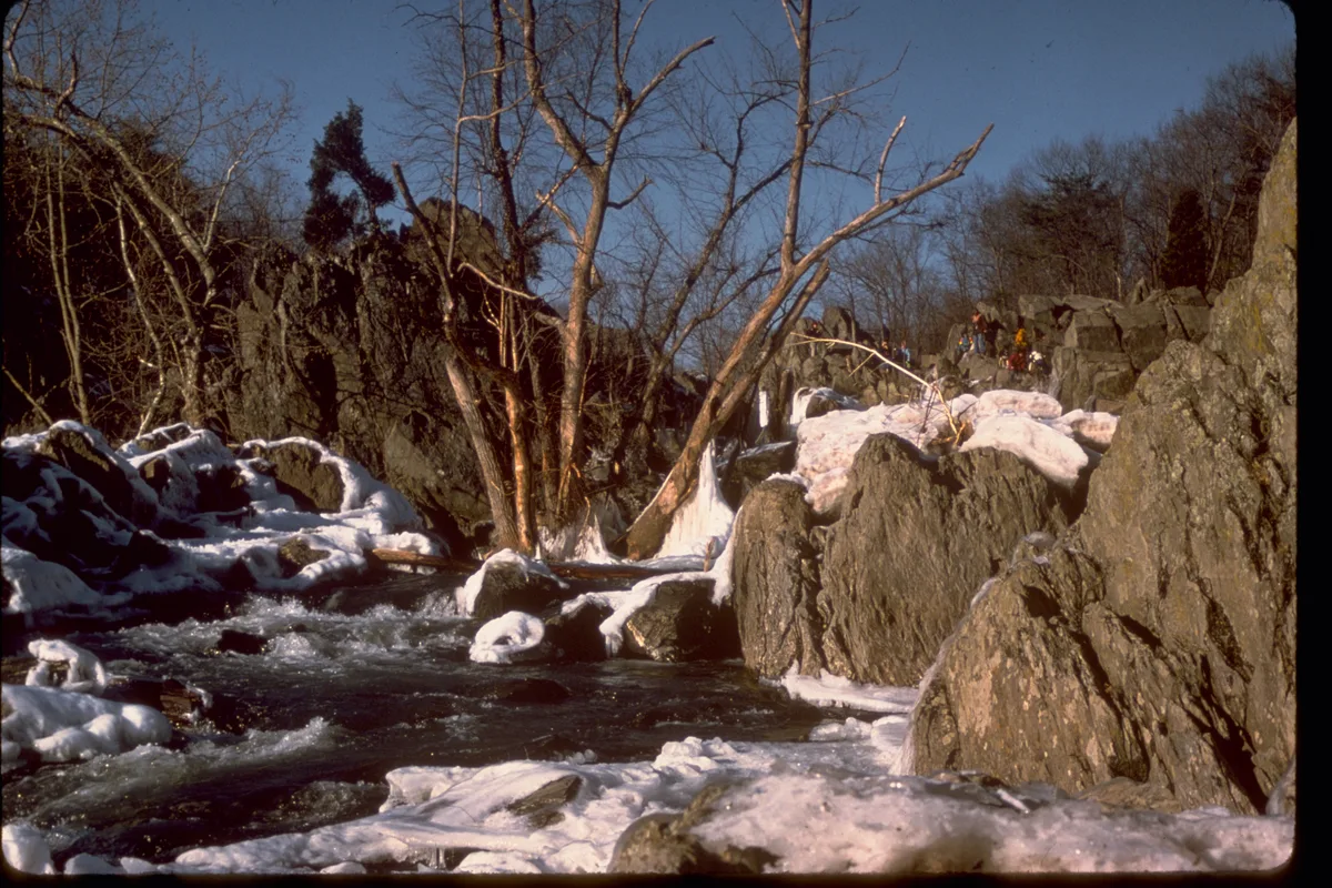 Sunlit winter rapids rush through a narrow rocky gorge at Great Falls, with snow and ice clinging to the boulders and leafless trees framing the scene. Visitors stand on the far overlook above the churning Potomac, under a clear blue sky.