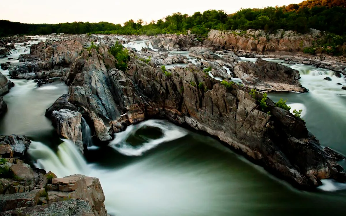 Wide view of the Potomac at Great Falls, with white water spilling through dark, jagged rock channels and around a narrow rocky island. Warm golden light touches the treeline along the horizon while the long-exposure water looks silky green and white.