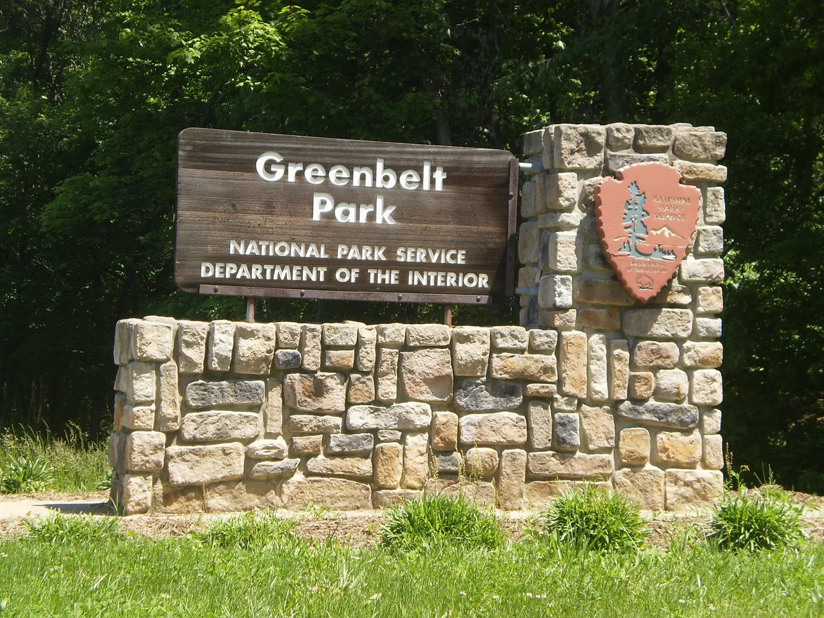 A stone and wood Greenbelt Park entrance sign stands in bright morning light, framed by dense green trees and fresh grass. The National Park Service arrowhead emblem is mounted on the right side of the stacked stone base.