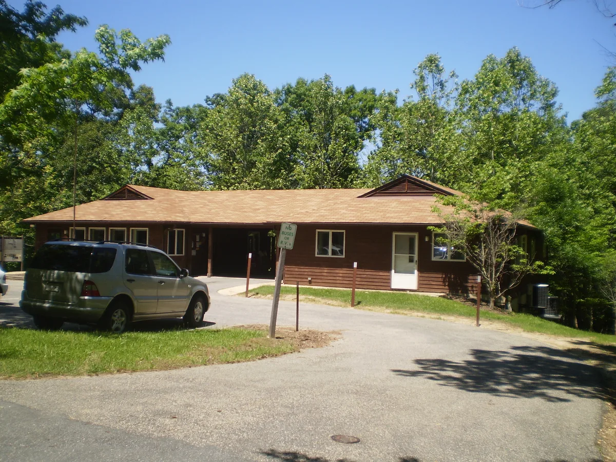 A low brown park building sits at the edge of a leafy forest clearing, with a silver SUV parked in front and a curved paved drive leading to the entrance. Bright morning light and a clear blue sky illuminate the dense green hardwood canopy behind it.
