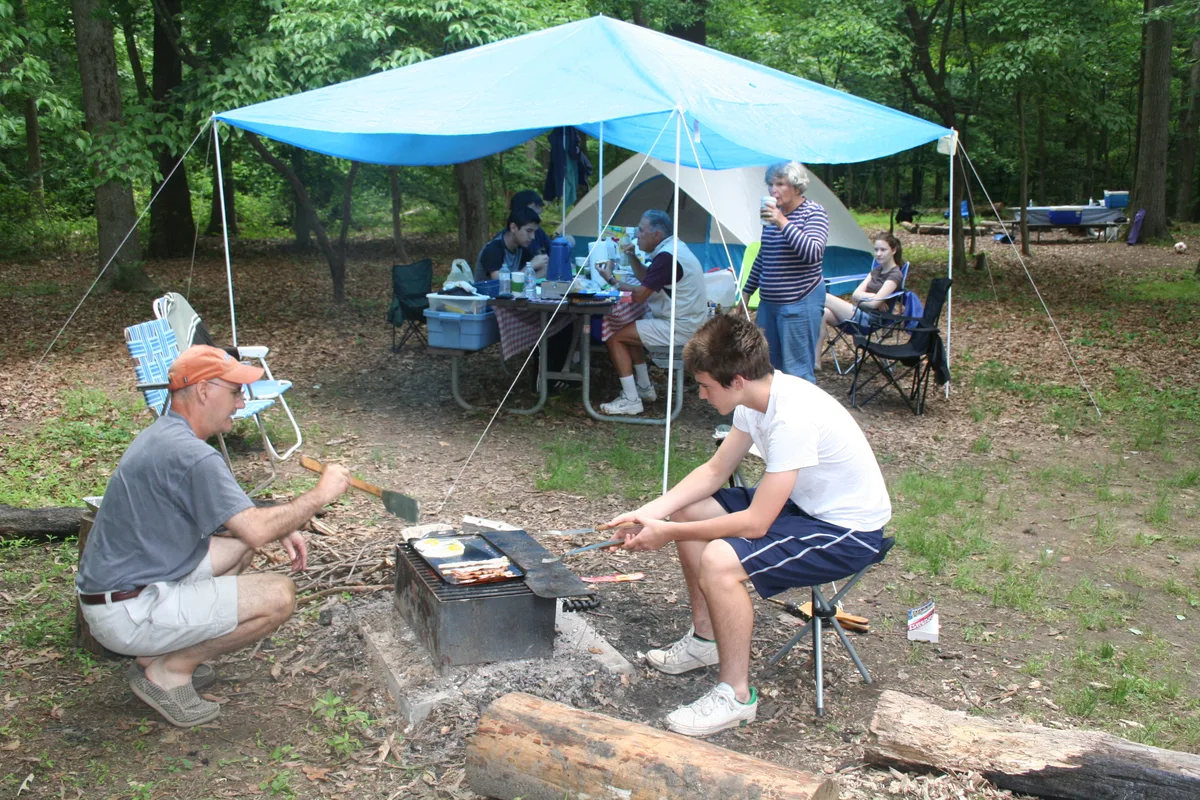 Campers gather in a wooded Greenbelt Park campsite, cooking breakfast over a small charcoal grill beneath a bright blue tarp. Foldable chairs, a tent, and scattered camping gear sit on the leaf-covered ground under tall green hardwood trees.