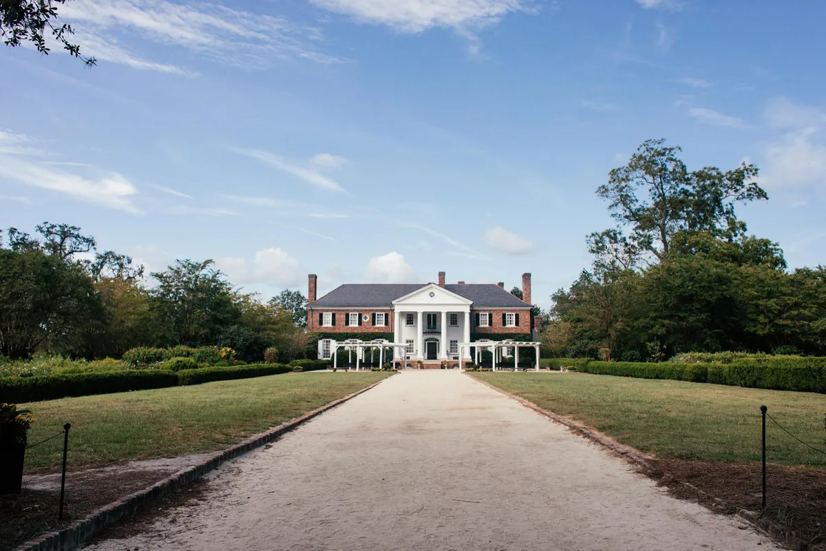 A long straight gravel path leads through clipped boxwood borders to Gunston Hall’s symmetrical colonial mansion, with a white-columned portico and red brick wings centered against a bright blue midday sky. Trees and low garden plantings frame the formal lawn on both sides, emphasizing the estate’s balanced, elegant layout.