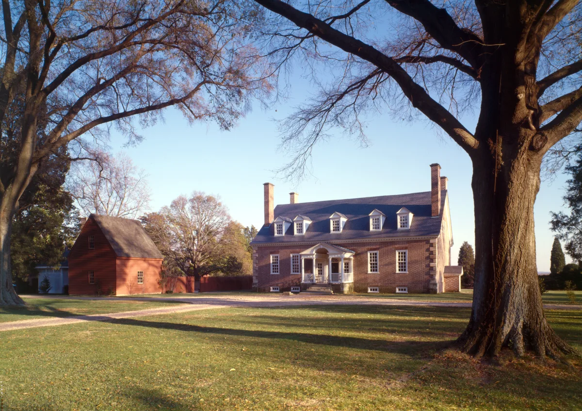 Gunston Hall’s brick Georgian manor sits in bright midday light, framed by two huge leafless trees and a wide lawn, with a red outbuilding to the left. The low, symmetrical house shows dormer windows, tall chimneys, and a small columned portico, set against bare branches and a pale blue sky.