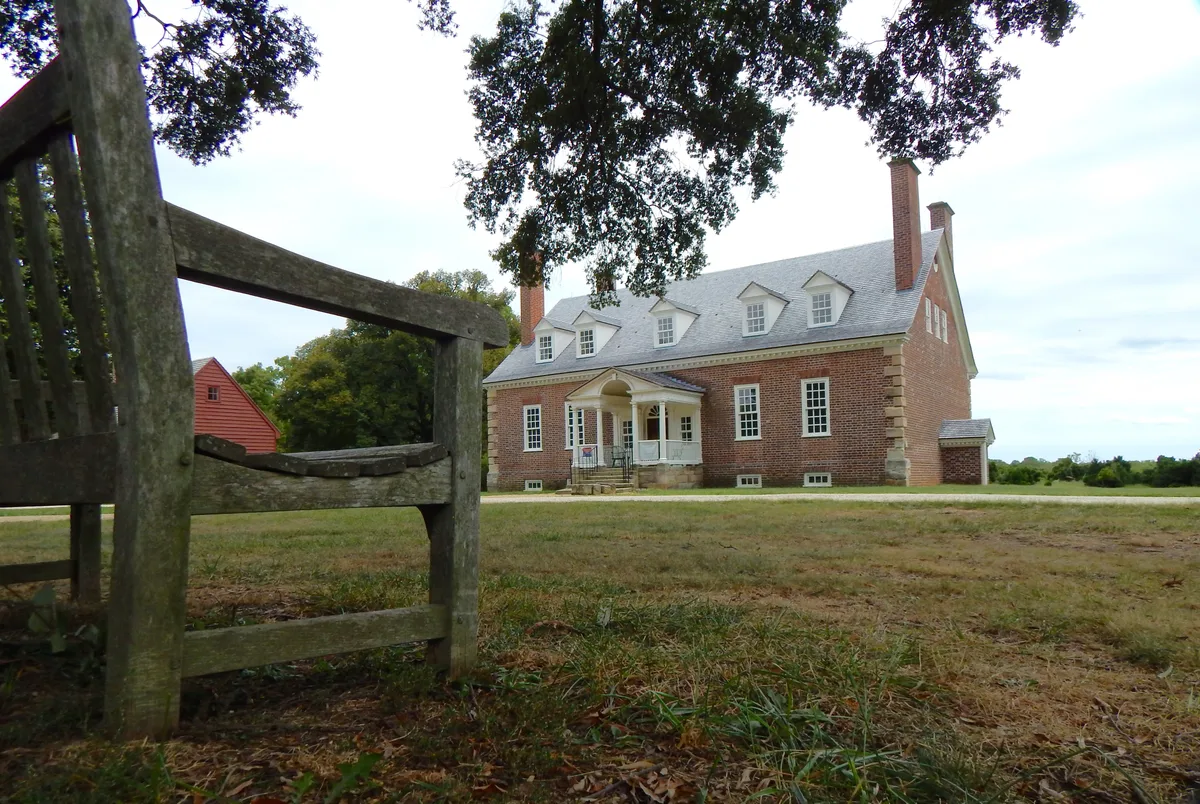A wide, low-angle view of Gunston Hall shows the red-brick colonial mansion centered in a broad grassy clearing, with a weathered wooden fence and overhanging tree branches framing the foreground. Bright midday light picks out the white porch, dormer windows, and tall chimneys against a pale sky.