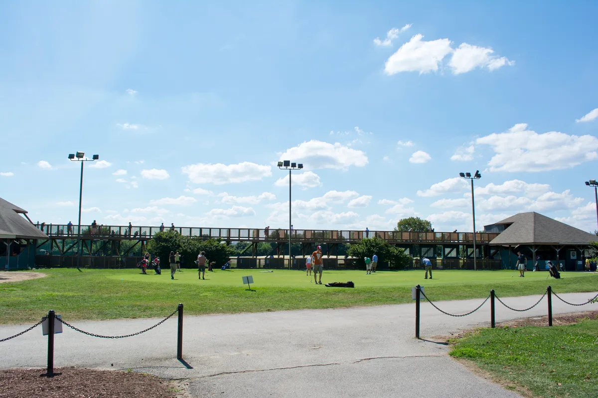 A wide view of the East Potomac Golf Course driving range at Hains Point, with golfers scattered across a bright green practice field under a blue sky with scattered clouds. A low wooden deck, tall light poles, and pavilion roofs frame the scene, giving it an open, park-like feel.