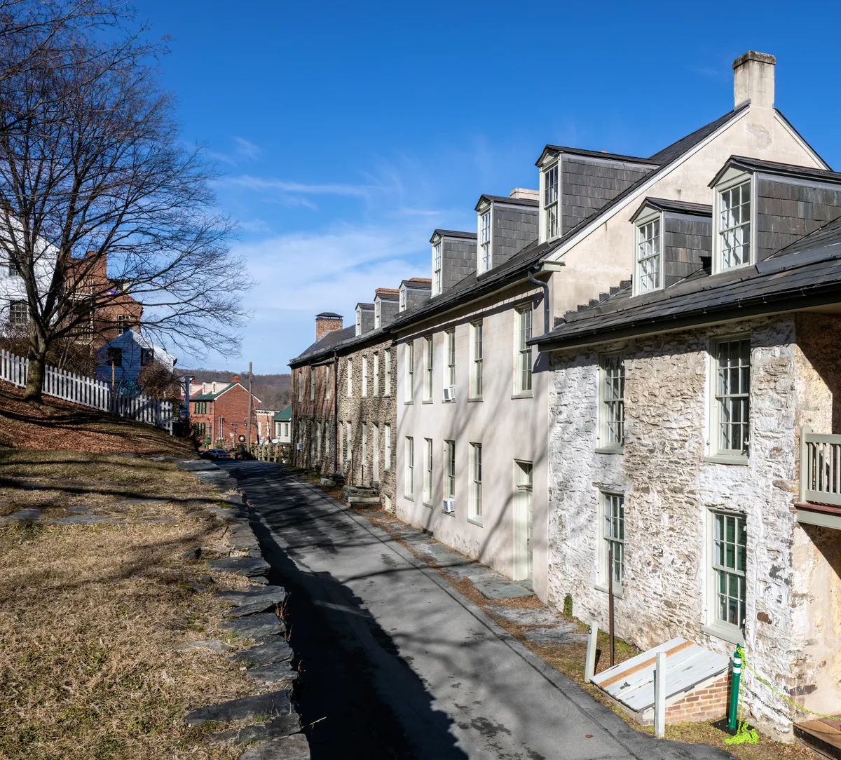 A narrow paved lane curves downhill between weathered stone and stucco historic row houses in Harpers Ferry, with dormer windows and chimneys catching bright sunlight under a clear blue sky. Bare winter trees and a white picket fence line the left side, emphasizing the town’s old, steep hillside setting.