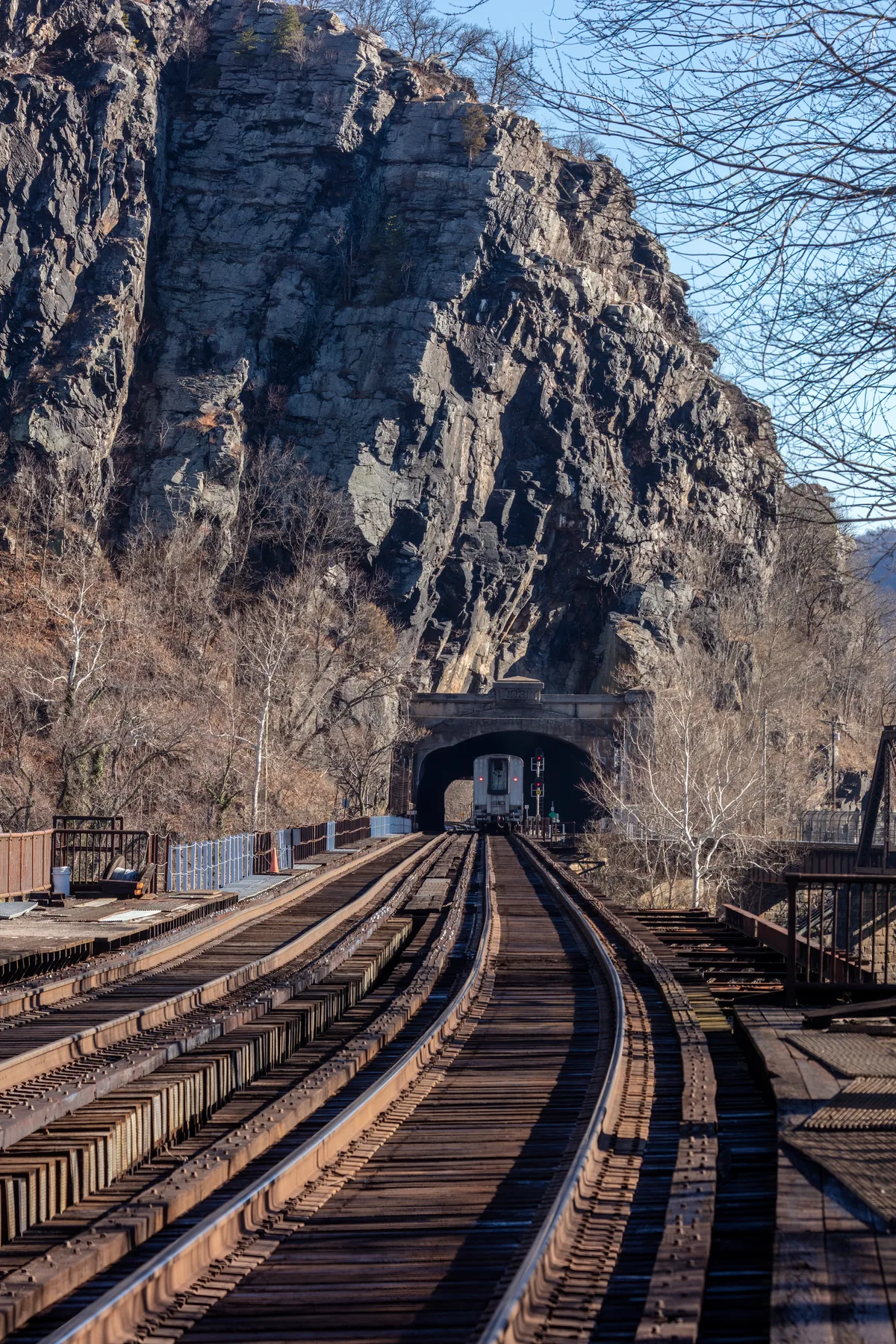 Railroad tracks curve toward a dark tunnel cut through towering gray cliffs at Harpers Ferry, with a train just emerging in the distance. Bare winter trees frame the scene, and the low golden light picks out the textures of the rock and weathered wooden trestles.