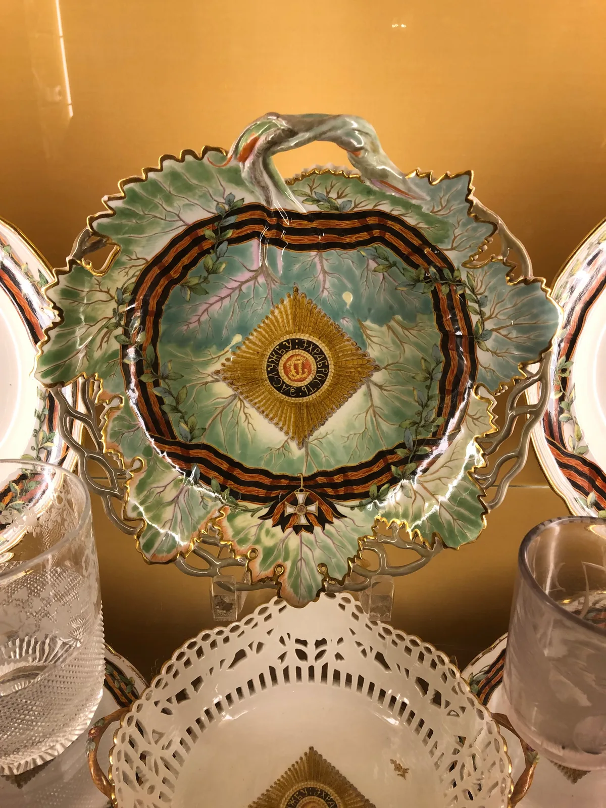 A gilded display cabinet holds ornate porcelain and glass, centered on a scalloped green leaf-shaped plate with gold edging and a dark ribbon motif. The warm amber background and close framing highlight the delicate painted details and the intricate cutout dish below.