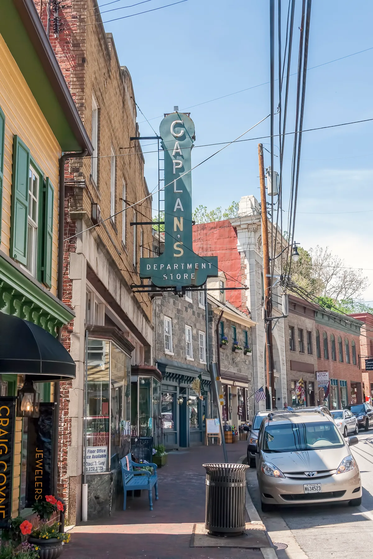 A narrow historic Main Street in Ellicott City is lined with old brick storefronts and hanging utility wires, dominated by a tall teal “Caplan’s Department Store” sign. Parked cars, sidewalk benches, and colorful shopfront details add to the small-town street scene in bright daylight.