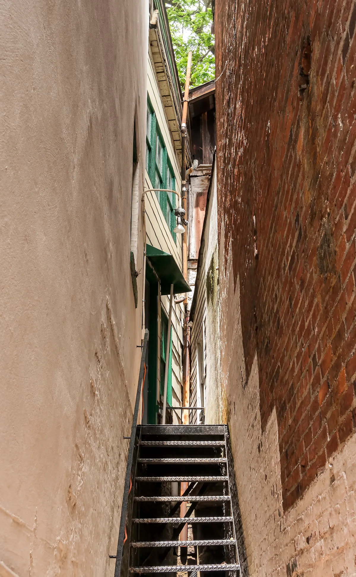 A narrow alleyway squeezes between a plain stucco wall and a weathered red brick wall, with a black metal staircase rising toward a tight gap of green trees overhead. Sunlight catches the worn surfaces, while a green-trimmed building and exposed pipes add to the old mill-town feel.