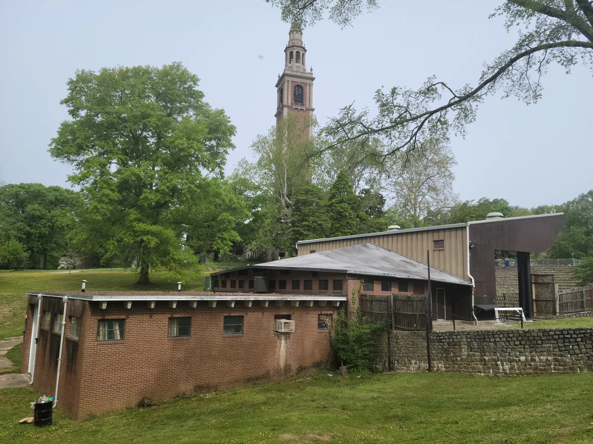 Brick and tan Pump House Park buildings sit in a leafy riverside setting, with a tall church steeple rising in the background above spring-green trees. The overcast light softens the scene, and a stone retaining wall and grassy slope frame the foreground.