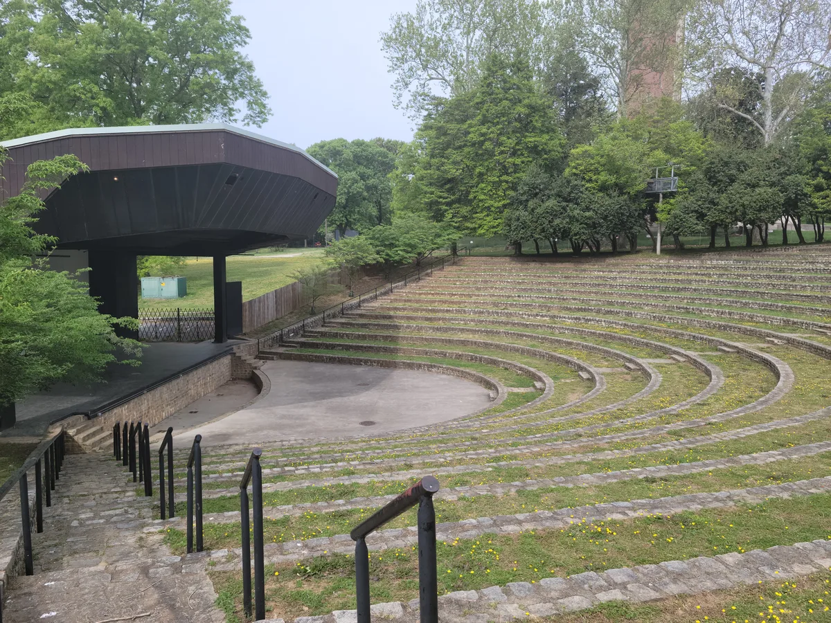 Curved stone amphitheater-style seating steps down toward a small stage beside a dark, angular pavilion, with black handrails leading through the foreground. Dense green trees and a brick chimney frame the park setting under soft, overcast light.