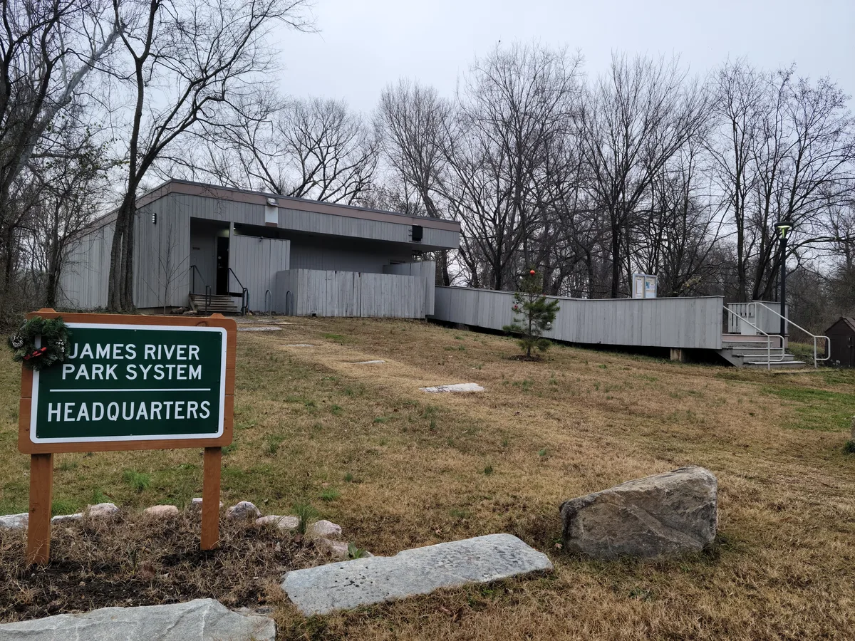 The James River Park System headquarters sits on a wintery hillside of brown grass and bare trees, with a green sign in the foreground and a simple gray building behind it. Concrete steps, a ramp, and scattered stones frame the quiet park setting under an overcast sky.