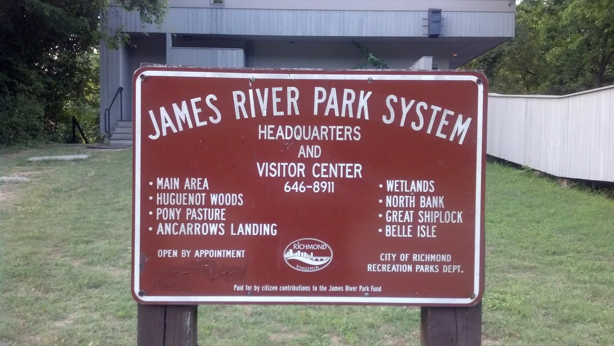 A brown sign for the James River Park System stands on a grassy patch in front of a low park building and trees. The sign lists the headquarters, visitor center, and park areas in white lettering, with a simple, utilitarian look.