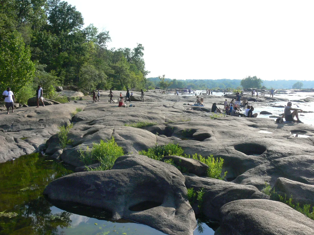People sit and walk across broad gray granite slabs along the James River at Belle Isle, with shallow pools in the foreground and whitewater rapids breaking to the right. Dense green trees line the left bank under bright, hazy light.