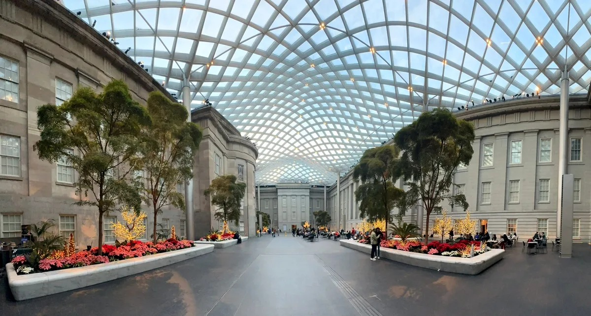 Wide view of Kogod Courtyard beneath a sweeping glass lattice roof, with soft midday light filtering through the canopy and casting a grid of pale shadows across the floor. Rectangular planters filled with red flowers and small trees line the museum facades, while visitors sit and walk along the open center aisle.