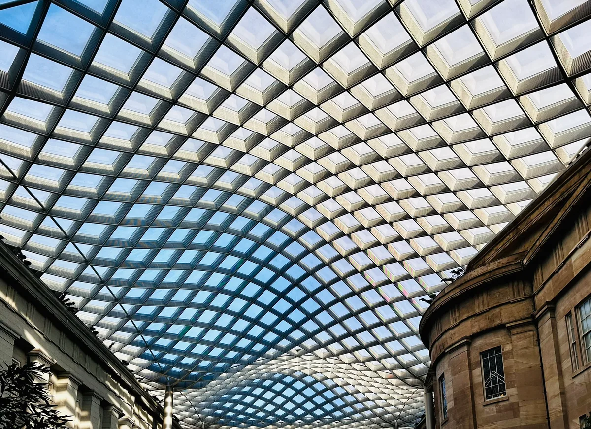 The Kogod Courtyard is covered by a sweeping diamond-pattern glass roof that curves overhead in pale blue and white bands of midday light. Historic stone walls frame the edges below, while the canopy creates a bold geometric abstract pattern across the ceiling.