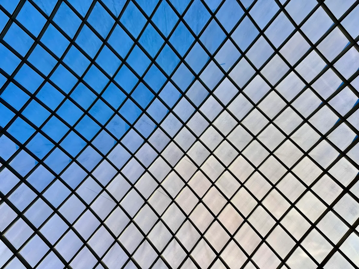 Looking straight up at Kogod Courtyard’s diamond-grid glass canopy, the black steel lattice forms a sharp geometric pattern against a blue sky streaked with pale clouds. The glass panels catch bright midday light, creating a cool, abstract ceiling effect.