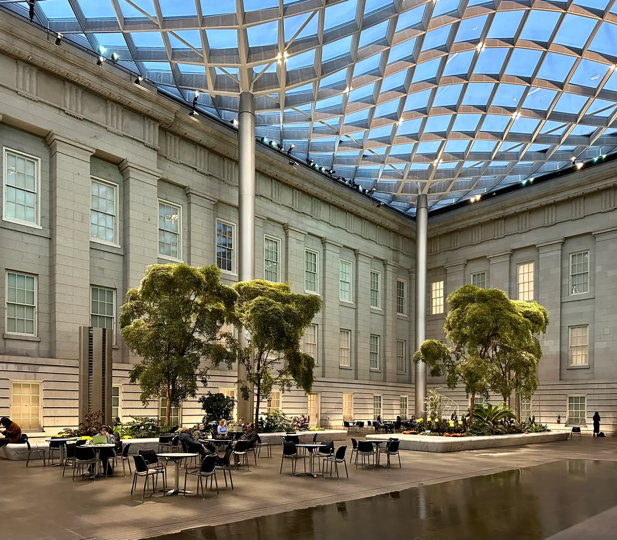 Kogod Courtyard’s glass lattice canopy arcs overhead, throwing bright geometric light across the museum’s pale stone walls and the open seating below. Small indoor trees and scattered café tables sit around a reflective floor, giving the space a calm, airy feel.