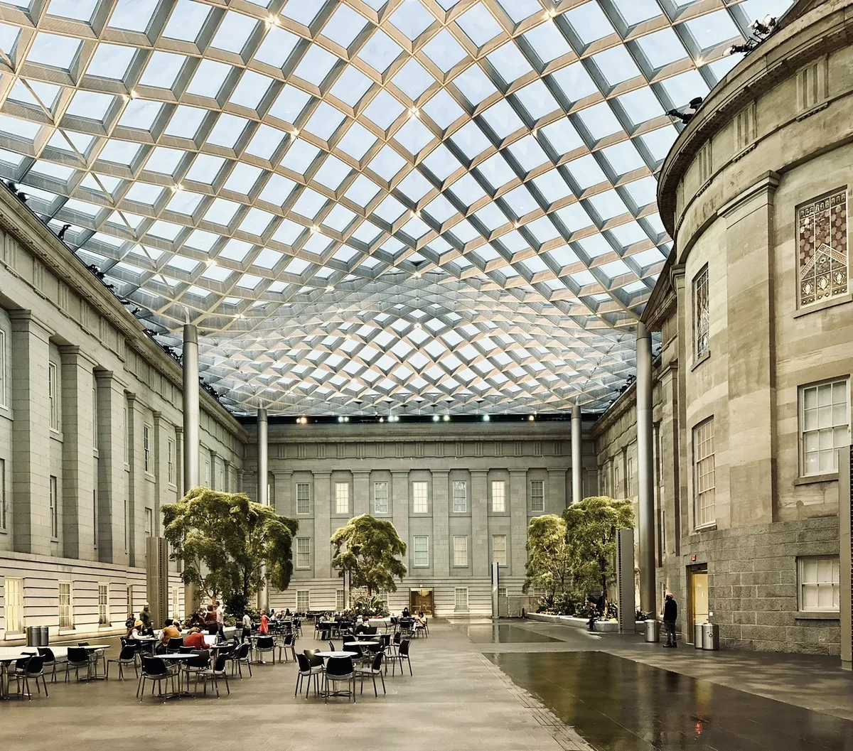Kogod Courtyard’s glass wave canopy fills the frame overhead, its diamond grid catching bright midday light and creating a luminous, geometric ceiling. Below, the stone museum walls curve around scattered café tables and small indoor trees, giving the space a calm, open-air feel.