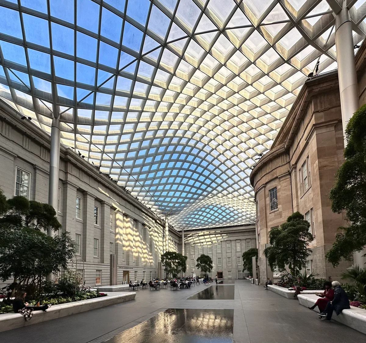 Kogod Courtyard’s sweeping glass roof ripples overhead in a blue-and-gold grid, casting bright geometric light and shadows across the museum’s pale stone walls. Below, benches, potted trees, and a few visitors sit around the open central space, with shallow reflective pools catching the ceiling’s pattern.