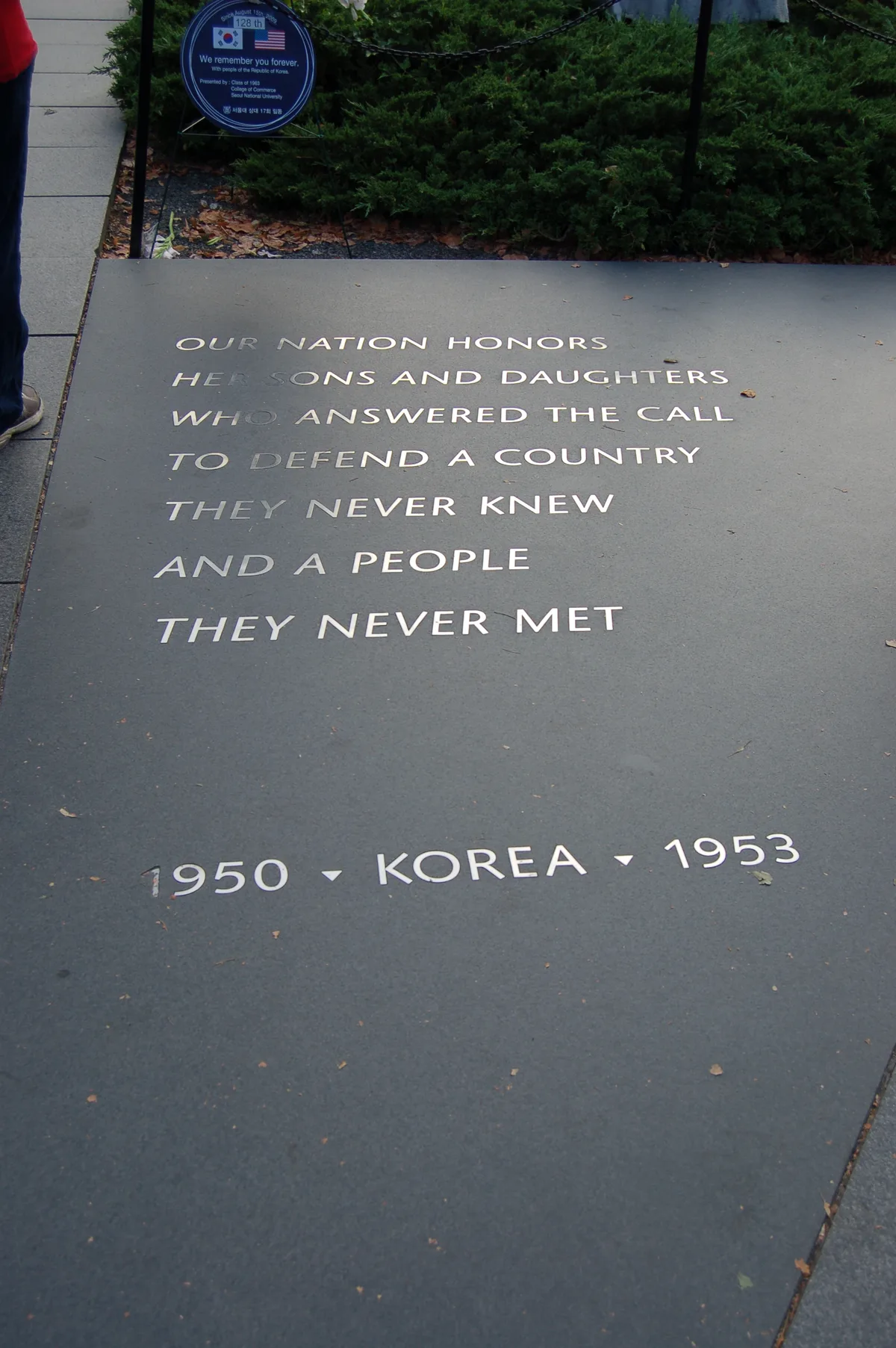 A close view of the Korean War Veterans Memorial’s dark granite walkway shows the etched dedication, “OUR NATION HONORS… THEY NEVER MET,” ending with “1950 • KOREA • 1953.” Juniper bushes and a small commemorative sign sit at the top edge, with a visitor’s legs partly visible along the left side.