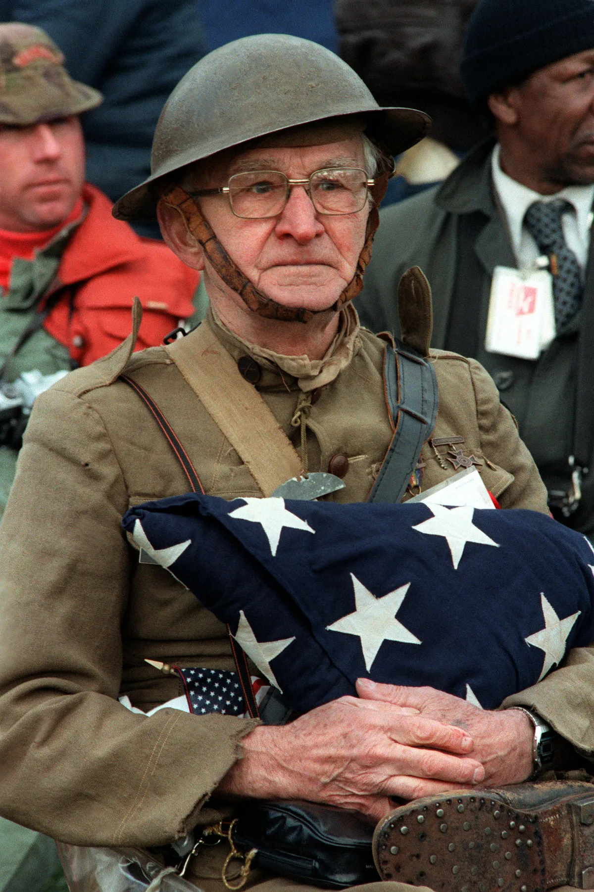 An elderly veteran in a khaki uniform and steel helmet sits solemnly in the foreground, clutching a neatly folded American flag across his chest. Other veterans blur behind him, adding to the quiet, commemorative mood.