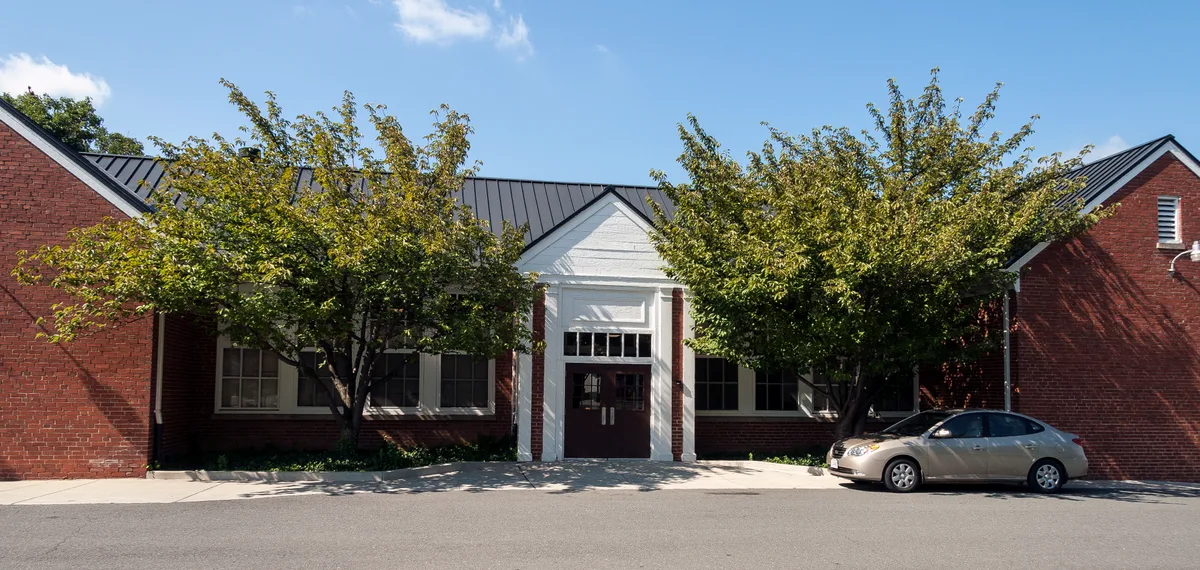 A low red-brick historic building sits in bright daylight, centered on a white-trimmed entrance with a black metal roof and flanked by leafy trees that cast soft shade across the facade. A silver car is parked at the curb in front, with a clear blue sky above.
