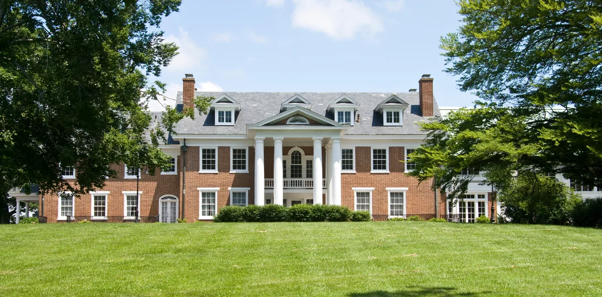 A grand red-brick colonial mansion sits behind a broad green lawn, framed by leafy trees and centered on a white-columned portico with a pediment and arched transom window. Bright midday light highlights the symmetry, dormer windows, and tall chimneys against a clear blue sky.