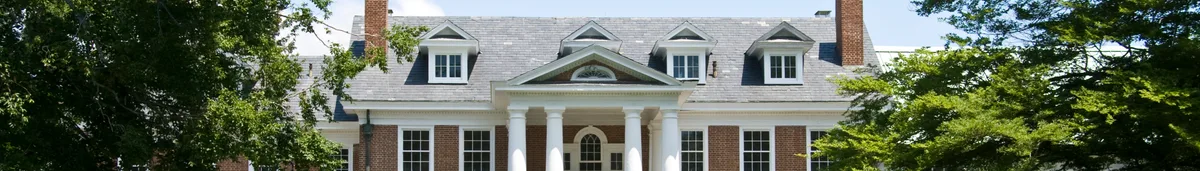 A stately red-brick colonial building with white columns, dormer windows, and a gray roof sits framed by leafy trees. The wide composition highlights the symmetry of the façade and the courthouse-like elegance of the historic streetscape.