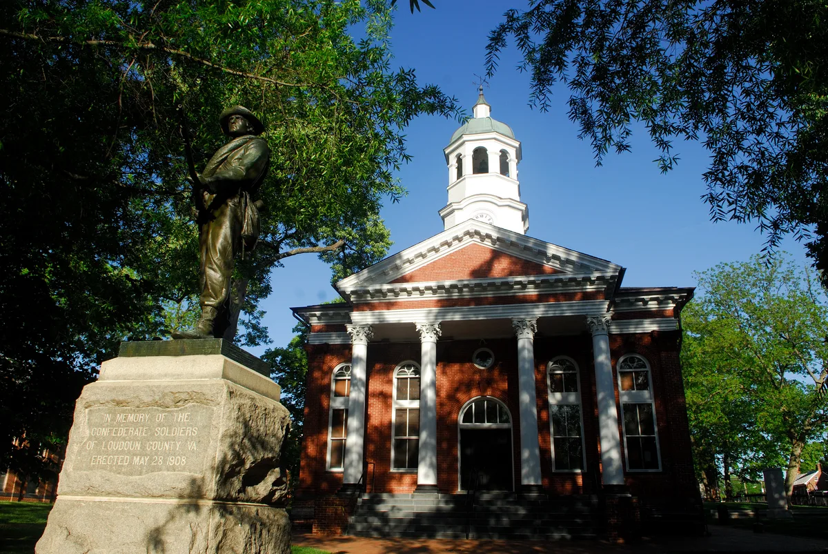 Sunlit Loudoun County Courthouse with a white columned portico and cupola, framed by leafy trees against a clear blue sky. A Confederate soldier statue on a stone pedestal stands in the foreground, its inscription visible in the warm late-day light.