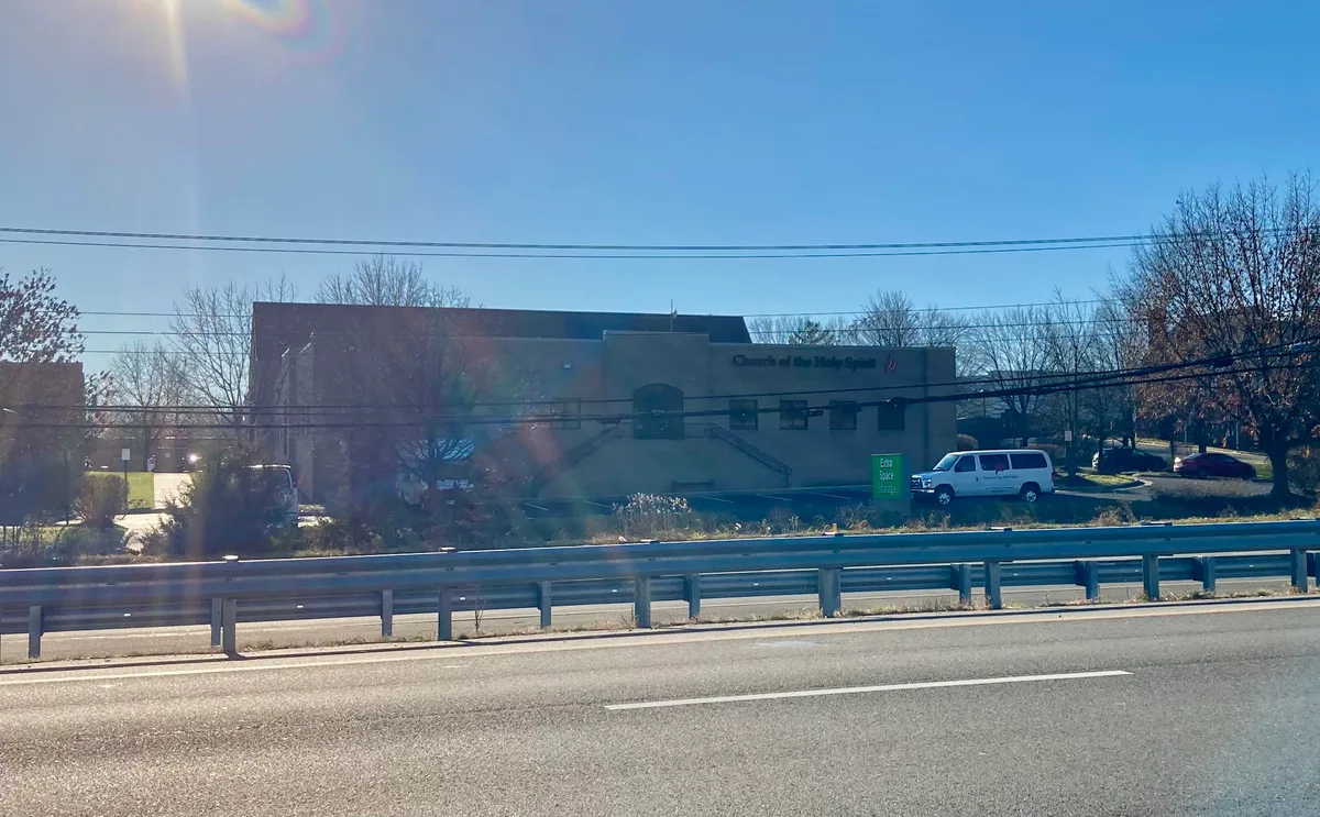 A low, beige church building sits back from the road behind a metal guardrail, with “Church of the Holy Spirit” visible on the facade and bare winter trees around it. Bright golden light and lens flare wash across the scene under a clear blue sky.