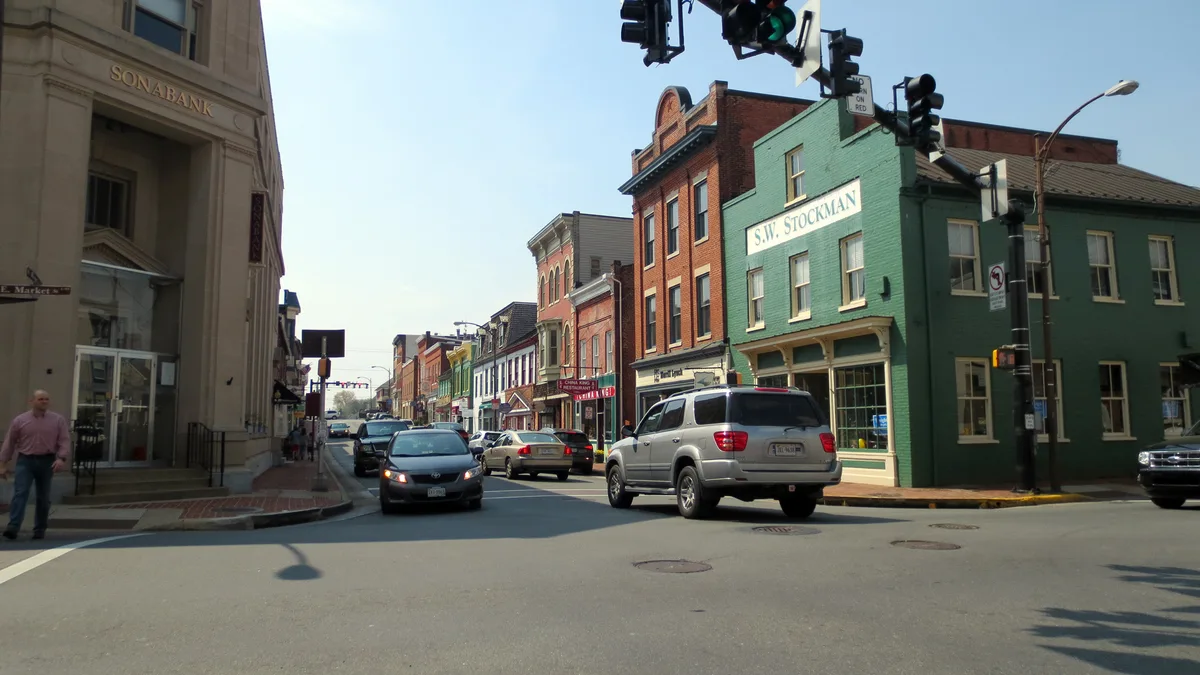 Historic brick storefronts line Leesburg’s main street, including the beige Sonabank building on the left and a green S.W. Stockman building on the corner, with cars moving through the intersection under traffic lights. Bright afternoon light washes the facades, highlighting the old-fashioned commercial streetscape.