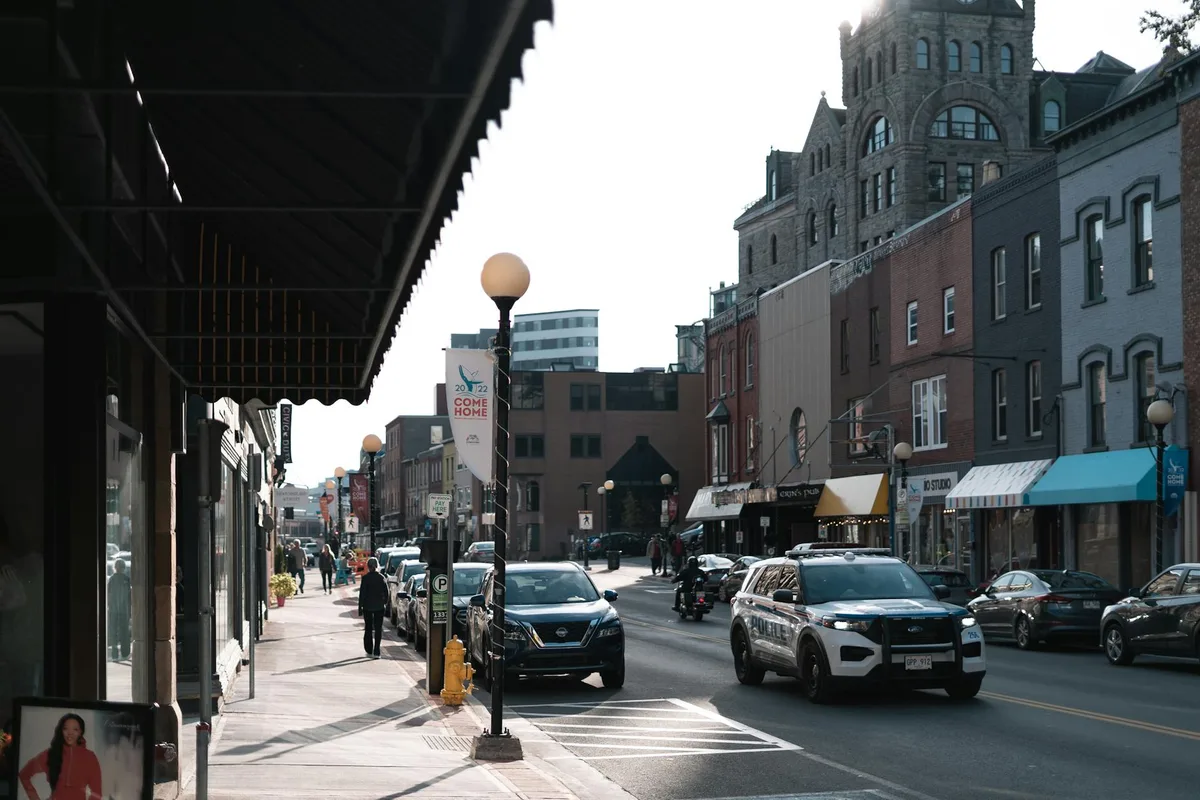 Golden-hour light washes over Leesburg’s brick-lined historic street, with storefront awnings, globe streetlamps, and a police SUV parked among cars along the sidewalk. A stone courthouse tower rises above the low-rise downtown buildings, adding a classic colonial streetscape feel.