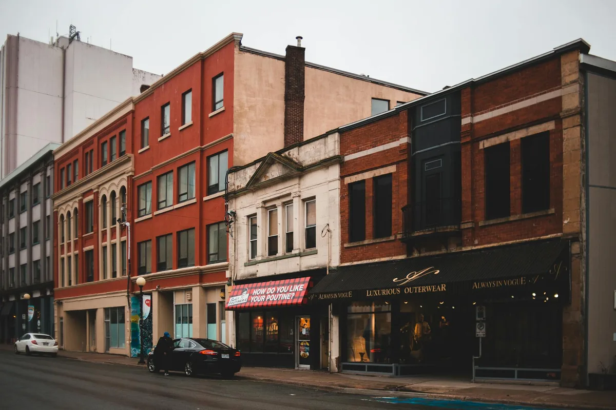 A row of mixed brick storefronts and upper-floor windows lines a quiet historic street, with a red-orange building, older white-trimmed facade, and dark awnings creating a layered streetscape. Parked cars and soft overcast light give the scene a muted, late-day feel.