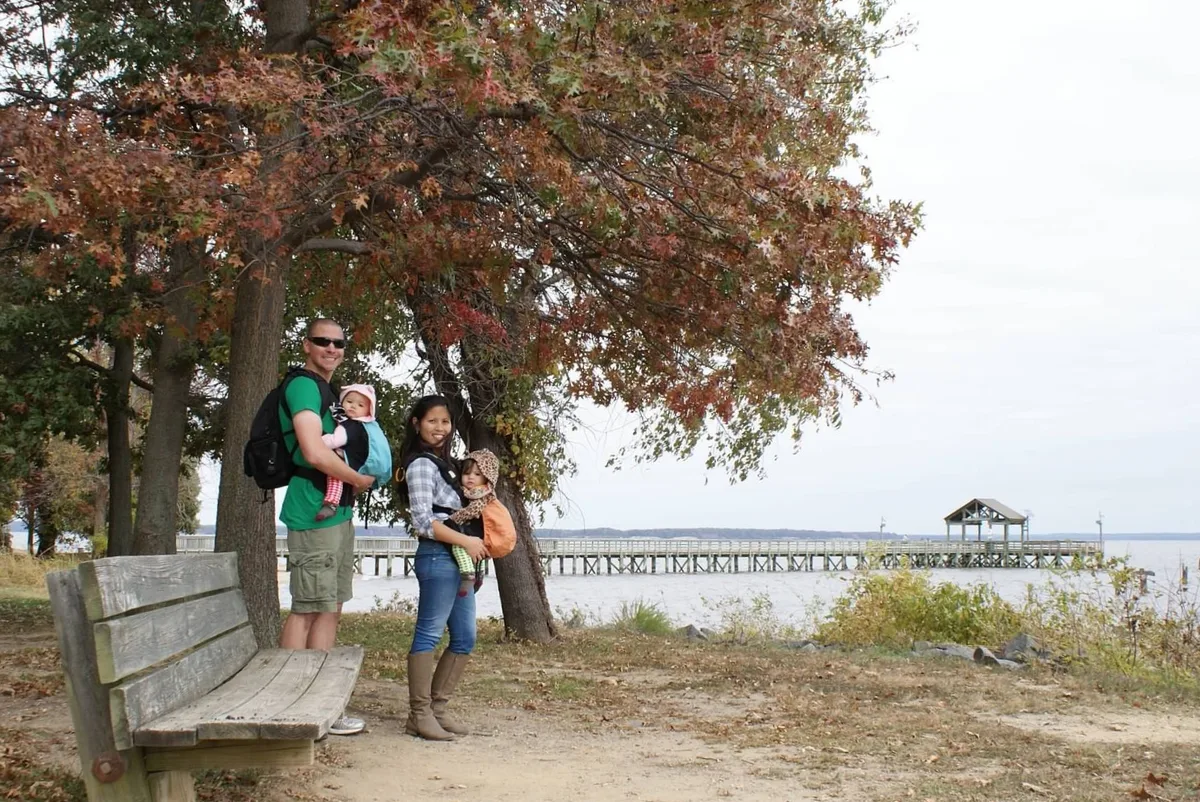 A family stands beneath autumn-colored trees beside a weathered wooden bench at Leesylvania State Park, with the Potomac River and a long pier stretching across the background. Soft overcast light brightens the wide shoreline view and distant water.