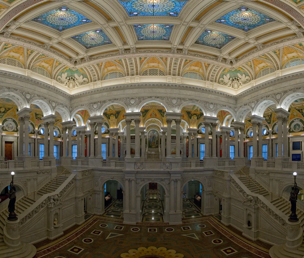 The Library of Congress Great Hall rises in pale marble with twin staircases, arched colonnades, and a richly patterned mosaic floor. Above, a vaulted ceiling glows with blue stained-glass panels, gilded ornament, and painted medallions that fill the space with midday light.