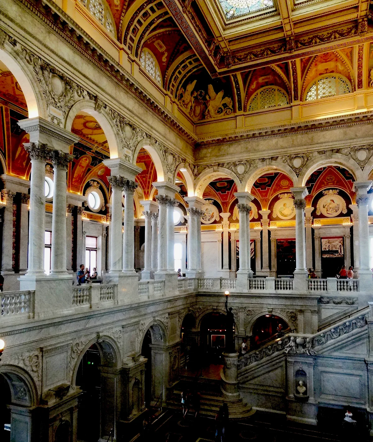 The Library of Congress Great Hall rises in layers of white marble arches and columns beneath a richly painted, gilded ceiling. Warm daylight filters through round and stained-glass windows, illuminating the mosaics, ornate friezes, and sweeping stairway below.