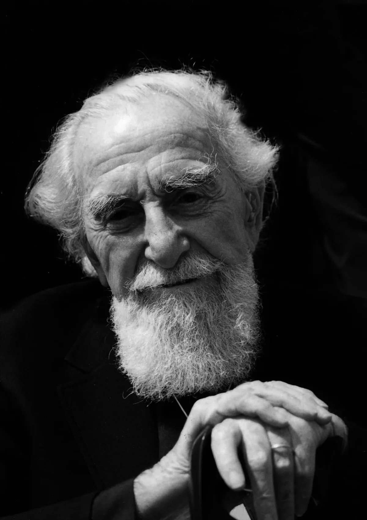 A black-and-white close-up portrait of an elderly man with wispy white hair, thick eyebrows, and a long full beard, posed against a dark background. His face is dramatically lit from the front, with his hands clasped over a cane in the lower foreground.
