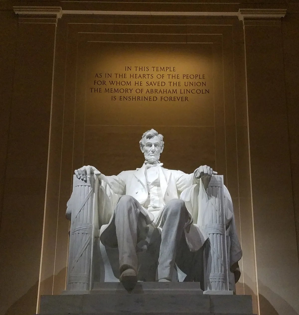 The seated marble statue of Abraham Lincoln fills the frame inside the memorial, lit warmly against a dim bronze wall. His name and the engraved dedication above him are clearly visible, emphasizing the solemn, monumental interior.